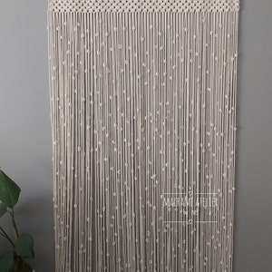 K&ouml;nnte beinhalten: Ein wei&szlig;er Makramee-Vorhang mit Perlenmuster. Der Vorhang ist an einer wei&szlig;en Stange aufgeh&auml;ngt und tr&auml;gt unten ein Etikett mit der Aufschrift "Macrame Atelier by Ines".
