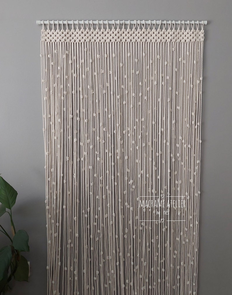 K&ouml;nnte beinhalten: Ein wei&szlig;er Makramee-Vorhang mit Perlenmuster. Der Vorhang ist an einer wei&szlig;en Stange aufgeh&auml;ngt und tr&auml;gt unten ein Etikett mit der Aufschrift "Macrame Atelier by Ines".