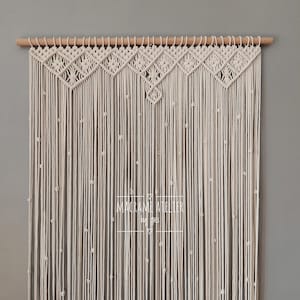 Può includere: Un arazzo da parete in macramè bianco con un motivo geometrico e una barra di legno nella parte superiore. L'arazzo è realizzato con cordoncino di cotone e ha una frangia nella parte inferiore. Il testo "MACRAME ATELIER by Ines" è visibile al centro dell'arazzo.