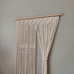 Könnte beinhalten: Ein weißer Makramee-Vorhang mit einer Holzstange oben. Der Vorhang besteht aus einem dicken, geknüpften Seil und hat ein geometrisches Muster oben. Der Text "MACRAME ATELIER" ist auf dem Vorhang sichtbar.
