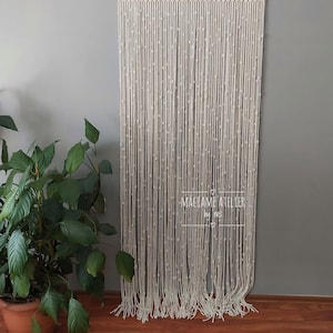 K&ouml;nnte beinhalten: Ein wei&szlig;er Makramee-Vorhang mit Perlenmuster. Der Vorhang h&auml;ngt an einer Wand und ist aus Naturfasern gefertigt. Der Text "MACRAME ATELIER by Ines" ist am unteren Rand des Vorhangs sichtbar.