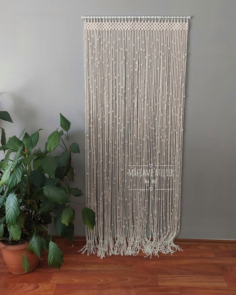 K&ouml;nnte beinhalten: Ein wei&szlig;er Makramee-Vorhang mit Perlenmuster. Der Vorhang h&auml;ngt an einer Wand und ist aus Naturfasern gefertigt. Der Text "MACRAME ATELIER by Ines" ist am unteren Rand des Vorhangs sichtbar.