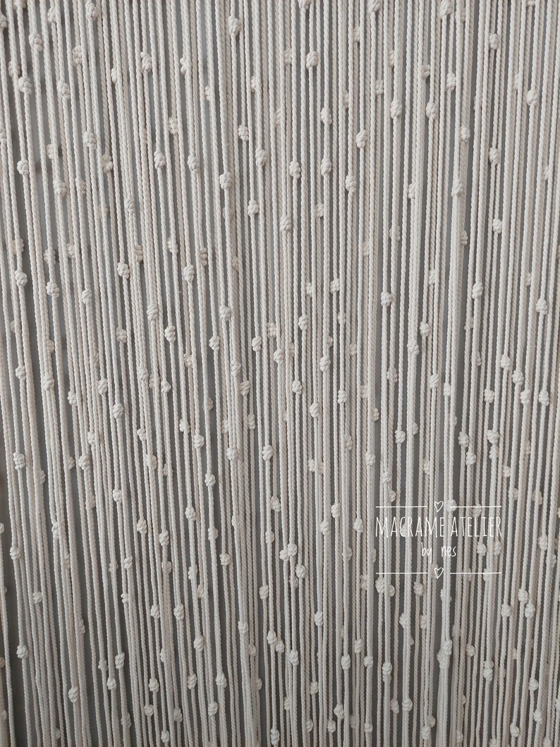 K&ouml;nnte beinhalten: Ein wei&szlig;er Makramee-Wandbehang mit einem sich wiederholenden Knotenmuster. Der Behang ist aus dickem Baumwollschnur gefertigt und hat eine subtile Textur. Der Text "MACRAME ATELIER" ist in der rechten unteren Ecke des Bildes sichtbar.