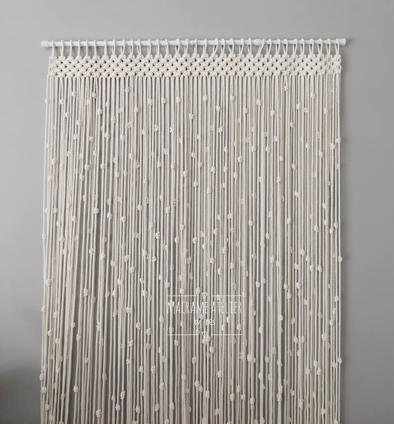 K&ouml;nnte beinhalten: Ein wei&szlig;er Makramee-Vorhang mit Perlenmuster. Der Vorhang ist an einer wei&szlig;en Stange aufgeh&auml;ngt und tr&auml;gt unten ein Etikett mit der Aufschrift "Macrame Atelier".