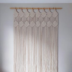 May include: A macrame wall hanging in a natural off-white hue. The design features diamond-shaped knots across the top, with long, vertical fringe below. It is suspended from a wooden rod with metal rings.