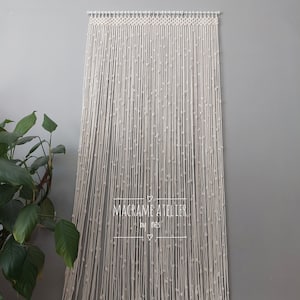 K&ouml;nnte beinhalten: Ein wei&szlig;er Makramee-Wandbehang mit Perlenmuster. Der Behang ist aus Baumwollschnur gefertigt und hat einen schlichten, minimalistischen Stil. Der Text "MACRAME ATELIER by nes" ist in der Mitte des Behangs sichtbar.