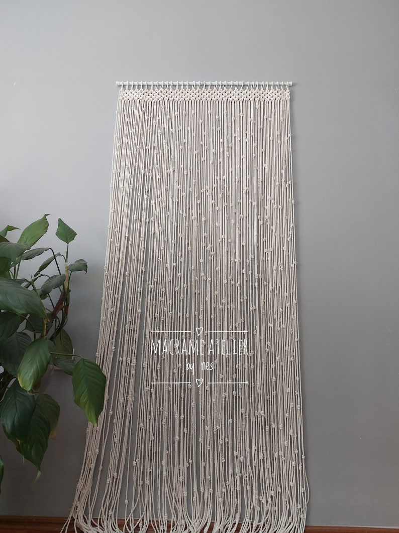 K&ouml;nnte beinhalten: Ein wei&szlig;er Makramee-Wandbehang mit Perlenmuster. Der Behang ist aus Baumwollschnur gefertigt und hat einen schlichten, minimalistischen Stil. Der Text "MACRAME ATELIER by nes" ist in der Mitte des Behangs sichtbar.