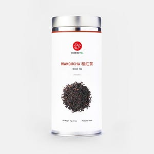 May include: A cylindrical white tea tin with a silver lid and a red band. The label features the text "WAKOUCHA Black Tea" in English and Japanese, a red floral logo, and a pile of loose tea leaves. Net weight is 2.6 oz.
