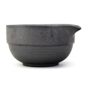 May include: A dark gray ceramic bowl with a spout. The bowl has a textured surface and horizontal ridged detailing around the base. The bowl is designed for pouring liquids.