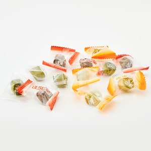 May include: A pile of individually wrapped green and brown candies in clear plastic bags with orange and red accents. The bags have Japanese text on them.