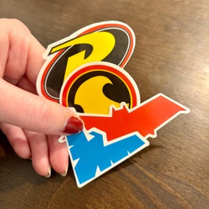 May include: A collection of three die-cut vinyl stickers featuring superhero-themed logos. One sticker displays a yellow and black R emblem, another shows a yellow and black circular bat symbol, and the third is a red and blue bat-shaped logo.