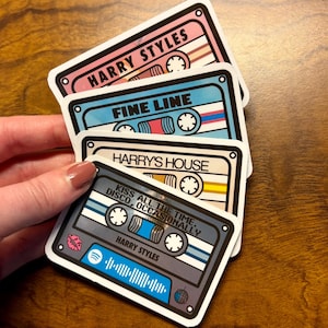 May include: A stack of four cassette tape-shaped stickers. The stickers feature the text "HARRY STYLES", "FINE LINE", "HARRY'S HOUSE", and "KISS ALL THE TIME, DISCO, OCCASIONALLY". The stickers have a retro aesthetic with colorful designs.