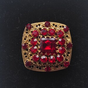 May include: A vintage square brooch with a gold filigree frame. The brooch features a large, square, red gemstone in the centre, surrounded by smaller, round red gemstones. The brooch is set against a black background.