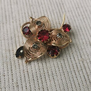 May include: A gold-toned floral brooch with red and gray gemstones. The brooch features a detailed design with swirling patterns and several red, round, and oval-shaped gemstones. The brooch is set against a textured, off-white background.