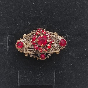 Op de afbeelding: Een broche in antieke stijl met een filigraan ontwerp. De broche heeft een cluster van levendige rode edelstenen gezet in een goudkleurig metalen frame. De edelstenen zijn van verschillende grootte, met een grotere centrale steen.