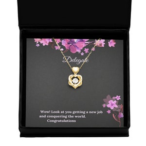 May include: A gold necklace with a spinning heart pendant and a clear stone in the center. The necklace is in a black box with a white card that says "Delegafe" and "Wow! Look at you getting a new job and conquering the world. Congratulations."