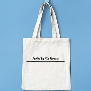 May include: A cream-coloured canvas tote bag with matching handles. The bag displays the black text "Fueled by Hip Thrusts" with a line beneath. It hangs against a light blue wall.
