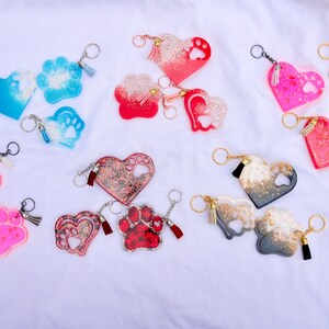 May include: A collection of heart and paw-shaped keychains in various colors, including blue, red, pink, and gray. Each keychain features a paw print design and a tassel, attached to a silver keyring.