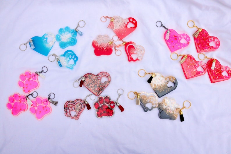 May include: A collection of heart and paw-shaped keychains in various colors, including blue, red, pink, and gray. Each keychain features a paw print design and a tassel, attached to a silver keyring.