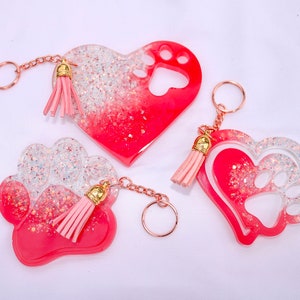 May include: Three resin keychains in heart and paw shapes. The keychains are red and clear with glitter accents and pink tassels. Each keychain has a gold-colored chain and ring.