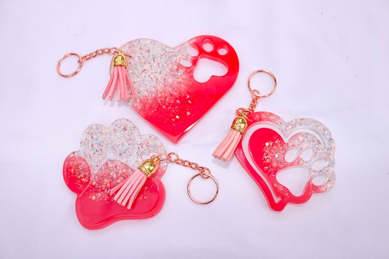 May include: Three resin keychains in heart and paw shapes. The keychains are red and clear with glitter accents and pink tassels. Each keychain has a gold-colored chain and ring.