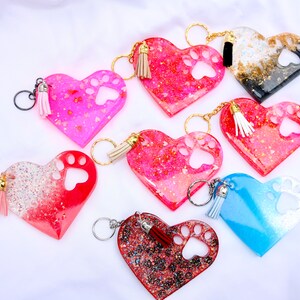 May include: Heart-shaped keychains in various colors, including pink, red, and blue, with paw print cutouts. Each keychain features a tassel and a gold or silver-colored keyring. The keychains are decorated with glitter and embedded accents.