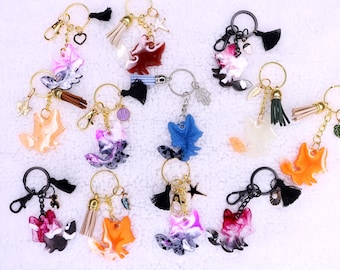 May include: A collection of fox-shaped keychains in various colors, including orange, pink, blue, and white. Each keychain has a metal ring, clasp, and decorative tassel or charm. Some charms include stars, leaves, and smiley faces.
