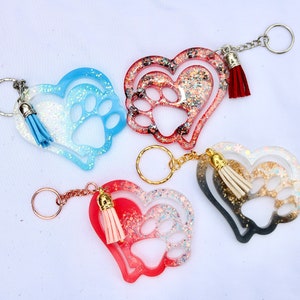 May include: Four heart-shaped keychains with paw prints and tassels. The keychains are in shades of blue, red, and black, with glitter accents. Each keychain has a metal keyring and a matching tassel.