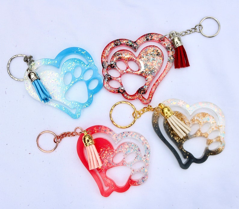 May include: Four heart-shaped keychains with paw prints and tassels. The keychains are in shades of blue, red, and black, with glitter accents. Each keychain has a metal keyring and a matching tassel.