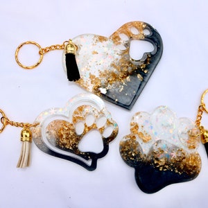 May include: Three resin keychains in heart and paw shapes. The keychains feature gold-colored accents, glitter, and black tassels. The heart-shaped keychains have paw print cutouts. The keychains are attached to gold-colored key rings.