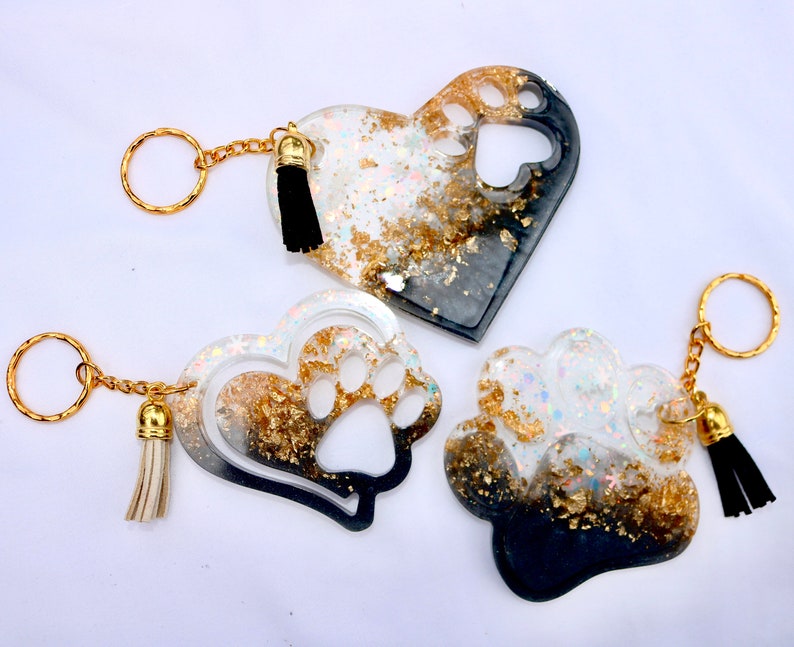 May include: Three resin keychains in heart and paw shapes. The keychains feature gold-colored accents, glitter, and black tassels. The heart-shaped keychains have paw print cutouts. The keychains are attached to gold-colored key rings.