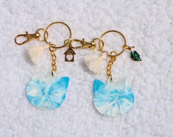 May include: Two cat-shaped keychains with gold-colored hardware. The cat pendants are blue and white with a marbled design. Each keychain has a tassel, a small charm, and a gold-colored clasp.