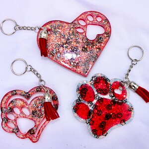 May include: Three glittery keychains in the shape of a heart and a paw print. The heart-shaped keychains have a paw print cutout. The paw print keychain is red with black paw prints. Each keychain has a silver chain and a red tassel.