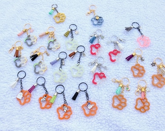 May include: A collection of keychains with various charms, including paw prints, ghosts, and abstract shapes. The keychains feature gold-colored hardware, colorful tassels, and small decorative charms. The keychains are in various colors, including orange, pink, and blue.