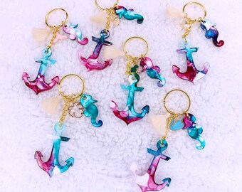 May include: A collection of anchor keychains with gold-colored rings. Each keychain features a colorful anchor pendant with blue, pink, and white hues. Some keychains include seahorse and seashell charms.