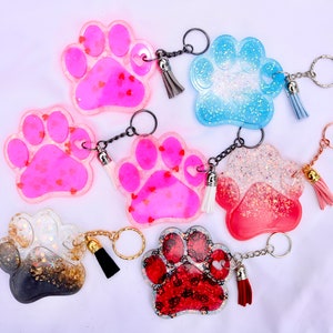 May include: A collection of paw-shaped keychains in various colors, including pink, blue, red, and black. Each keychain features a glittery design and a small tassel. Some keychains have heart-shaped accents.