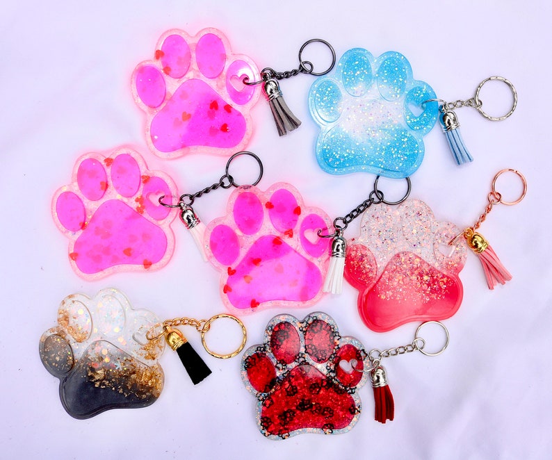May include: A collection of paw-shaped keychains in various colors, including pink, blue, red, and black. Each keychain features a glittery design and a small tassel. Some keychains have heart-shaped accents.
