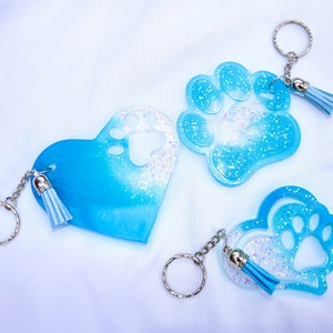May include: Three resin keychains in the shape of a heart, paw print, and heart with a paw print cutout. The keychains are blue with glitter and a silver keyring and tassel. The keychains are a pet-themed accessory.