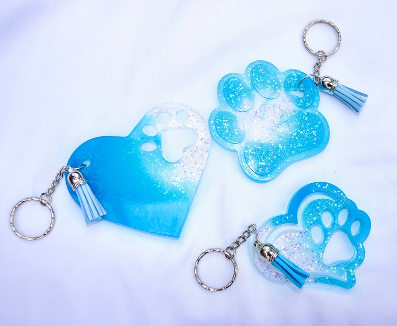 May include: Three resin keychains in the shape of a heart, paw print, and heart with a paw print cutout. The keychains are blue with glitter and a silver keyring and tassel. The keychains are a pet-themed accessory.