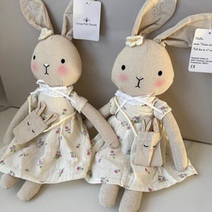 Könnte beinhalten: Zwei beigefarbene Hasenpuppen, die Blumenkleider tragen und kleine Hasentaschen mit sich führen. Die Puppen sind 47 cm groß und haben gestickte Buchstaben an den Ohren. Das Etikett lautet "Hallo, mein Name ist... Ich bin in 47 cm".