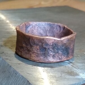 May include: A wide copper ring with a hammered texture. The ring is sitting on a metal surface.