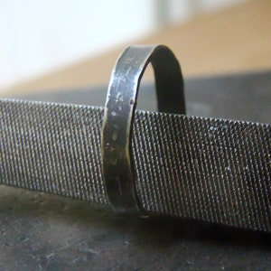 Puede incluir: Primer plano de un anillo de metal oscuro sobre una lima metálica. El anillo tiene una superficie texturizada, y la lima tiene una textura rugosa y estriada. El fondo está borroso, sugiriendo un taller.