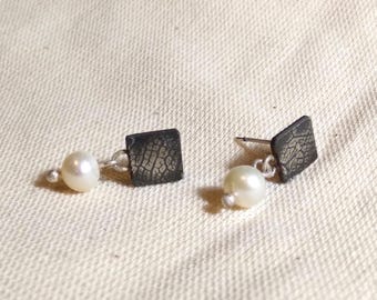 Op de afbeelding: Een paar oorbellen met een zwarte vierkante hanger en een witte parel die aan elke hanger hangt. De vierkante hanger heeft een getextureerd oppervlak.