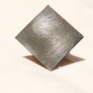 May include: A square, silver-colored metal tile with a brushed finish.
