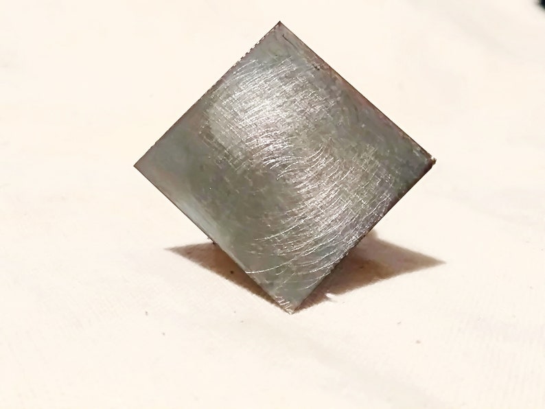 May include: A square, silver-colored metal tile with a brushed finish.
