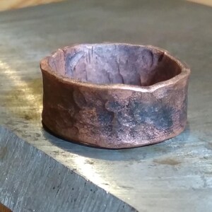 May include: A wide band ring crafted from copper with a dark, oxidized finish. The surface features a heavily hammered, irregular texture with visible indentations and a rugged, uneven edge along the top rim of the circular metal band.