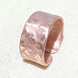 May include: A wide, hammered copper ring with a slightly textured finish. The ring is a warm, rose gold colour.