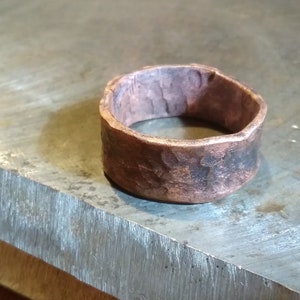 May include: A wide copper ring with a hammered texture. The ring is sitting on a metal surface.
