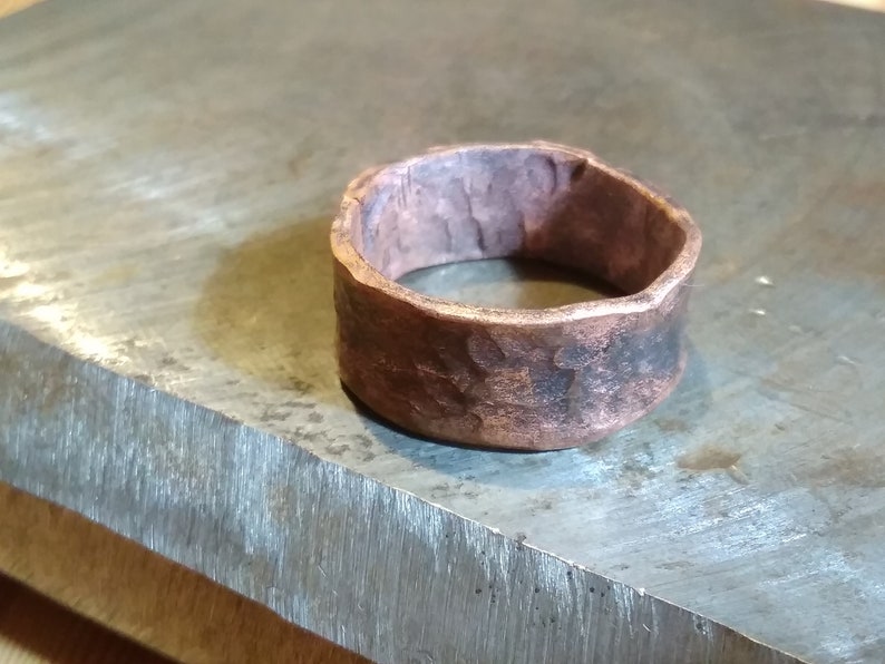 May include: A wide copper ring with a hammered texture. The ring is sitting on a metal surface.