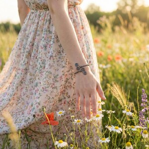 May include: A bronze-colored bracelet with spiral designs is worn on an arm. The person is wearing a floral print dress and is in a field of wildflowers. The bracelet is the focal point of the image.